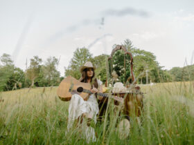 Country singer-songwriter Kylie Frey sitting in a field with her guitar and cowboy hat, promotional photo for her song “My John Wayne.”