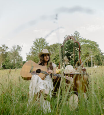 Country singer-songwriter Kylie Frey sitting in a field with her guitar and cowboy hat, promotional photo for her song “My John Wayne.”