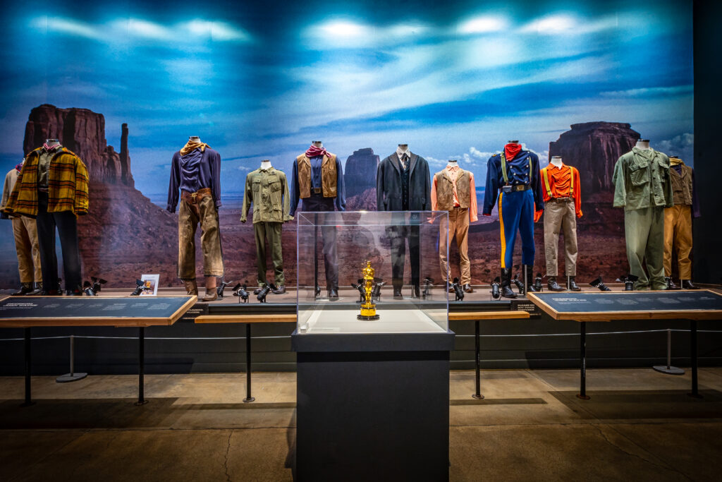 Display of original John Wayne film costumes arranged by era, with an Academy Award statue at center, inside the John Wayne: An American Experience museum in the Fort Worth Stockyards.